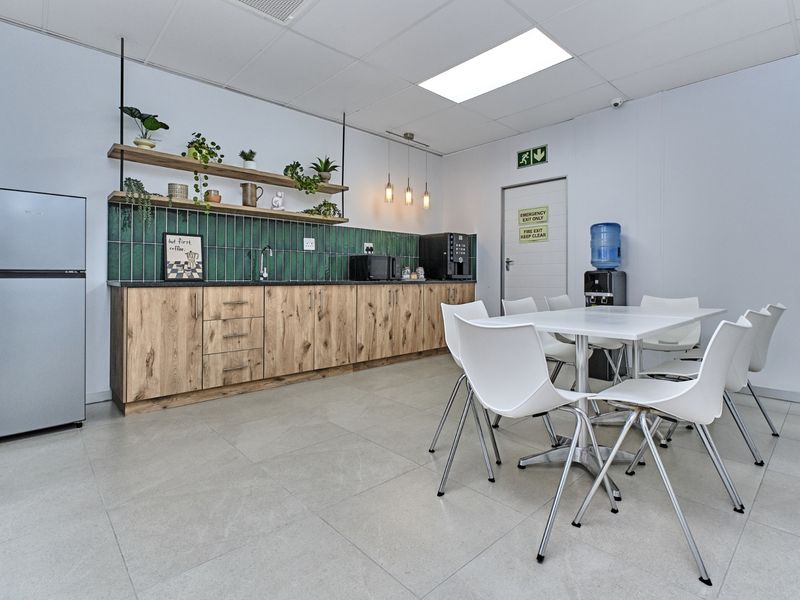 A modern commercial kitchenette featuring wood-grain cabinetry, a green tiled backsplash, floating shelving, and a dedicated dining area with tiled flooring.