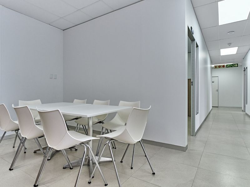 A clean, modern office meeting space featuring tiled flooring, suspended ceilings with recessed lighting, and access to a connecting hallway.