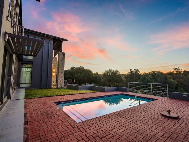 A well-maintained outdoor pool area featuring brick paving, stone accents, and a metal safety railing, set against a scenic sunset backdrop.