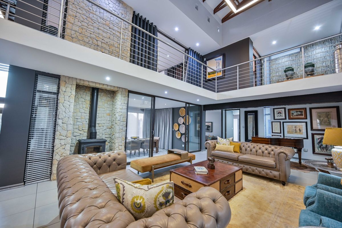 Spacious double-volume living area featuring a stone fireplace, mezzanine level with stainless steel balustrades, exposed timber rafters, and large sliding doors to the patio.