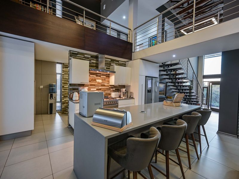 Modern open-plan kitchen featuring a large stone island, high-end built-in appliances, and a striking double-volume ceiling with a mezzanine level and feature staircase.