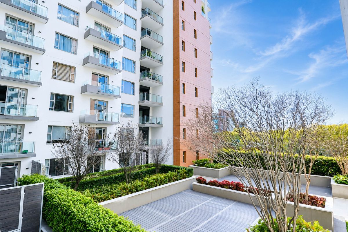 A view of the apartment building exterior featuring a landscaped terrace with raised garden beds, decking, and modern glass-balustraded balconies.