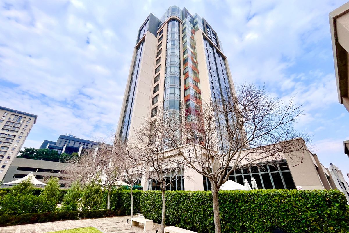A modern high-rise apartment building featuring a glass facade, set within a well-maintained landscaped garden area with stone pathways.