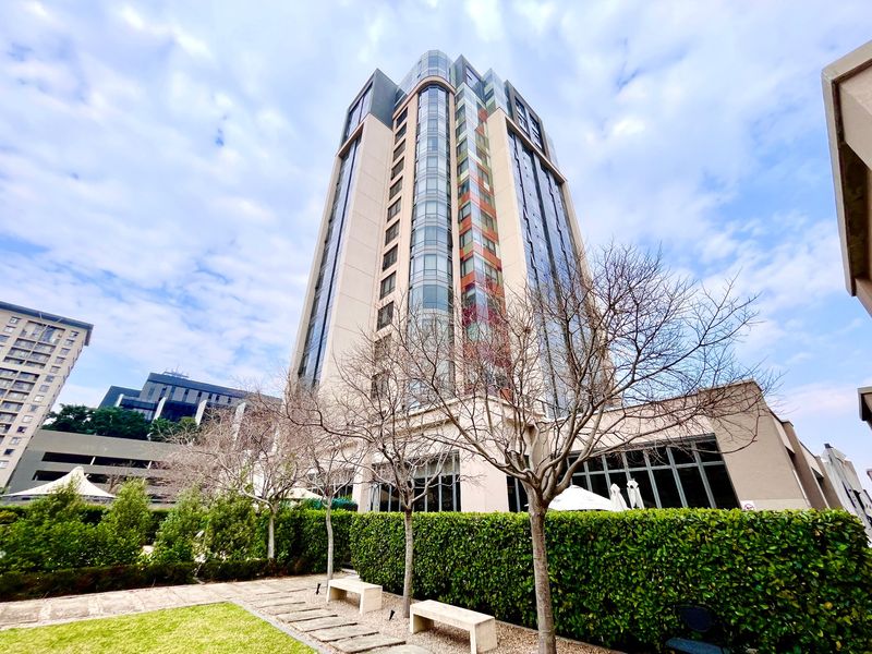 A modern high-rise apartment building featuring a glass facade, set within a well-maintained landscaped garden area with stone pathways.