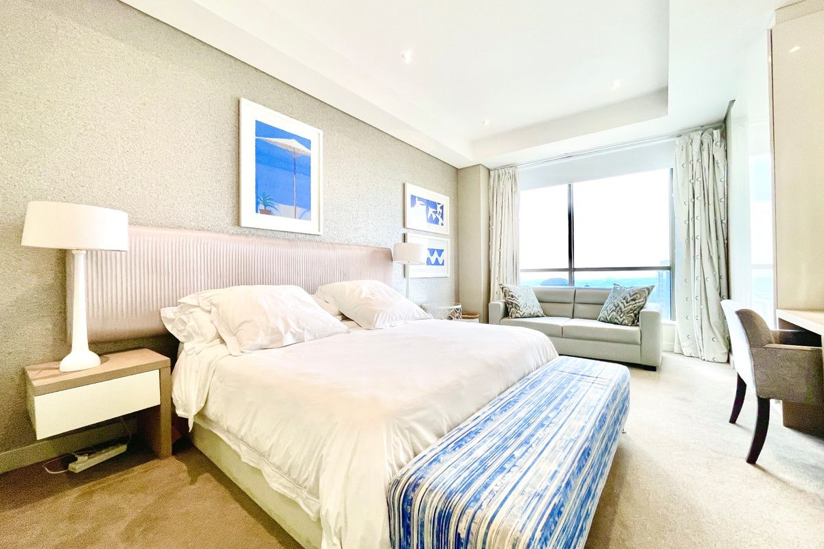 A well-maintained bedroom featuring a built-in headboard, recessed ceiling lighting, and large floor-to-ceiling windows offering natural light and views.