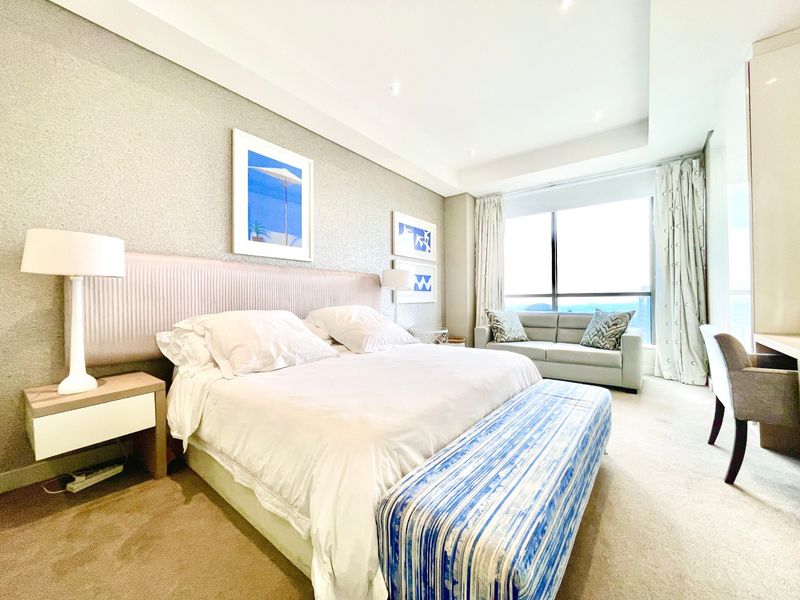 A well-maintained bedroom featuring a built-in headboard, recessed ceiling lighting, and large floor-to-ceiling windows offering natural light and views.