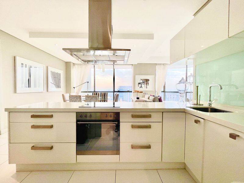 Modern open-plan kitchen featuring a central island, integrated stainless steel appliances, sleek cabinetry, and a glass splashback, flowing into a bright living area.