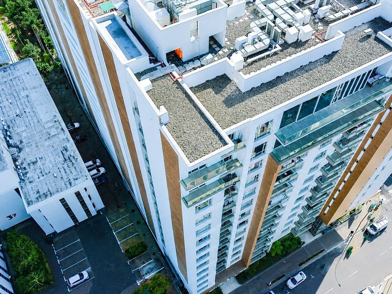 Aerial view of a modern multi-story apartment building featuring glass-fronted balconies, roof-mounted mechanical infrastructure, and an adjacent paved parking area.