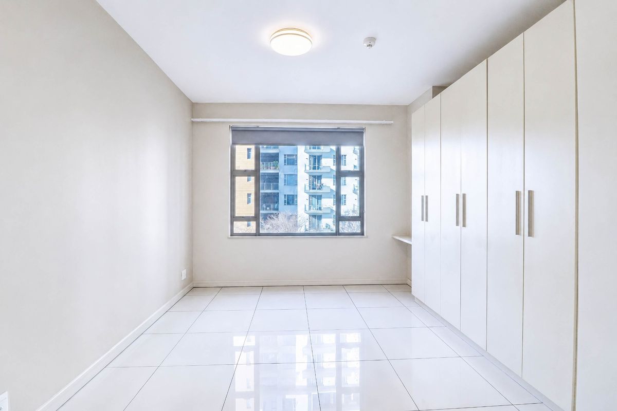 A bright, empty bedroom featuring neutral tiled flooring and extensive built-in cupboard storage with a window overlooking the surrounding apartment complex.