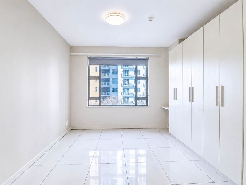 A bright, empty bedroom featuring neutral tiled flooring and extensive built-in cupboard storage with a window overlooking the surrounding apartment complex.