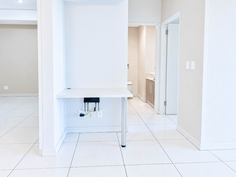 A bright, neutral hallway featuring white tiled flooring, a built-in desk area, and access to adjacent rooms.