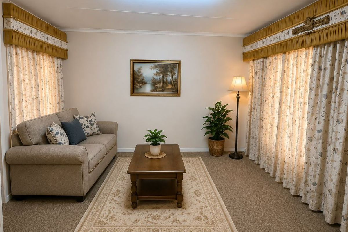 A carpeted living room featuring neutral walls, large windows with decorative curtain pelmets, and a central fluorescent light fixture.
