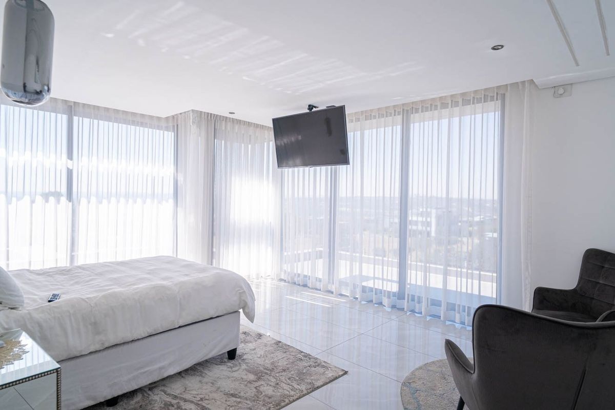 Spacious modern bedroom featuring floor-to-ceiling windows with sheer curtains, tiled flooring, and a wall-mounted television, offering a bright and contemporary living space.