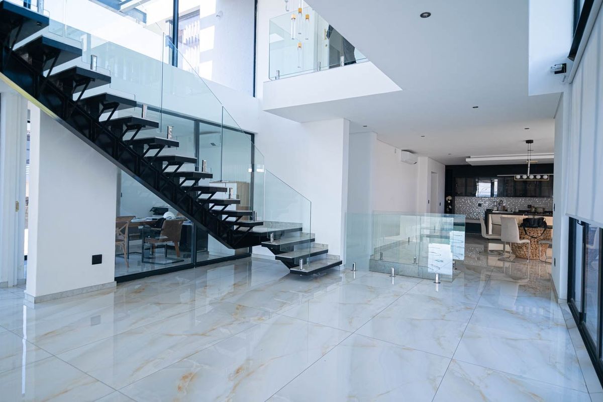 Spacious open-plan living area featuring a striking floating staircase with glass balustrades, high ceilings, and premium marble-look floor tiles.