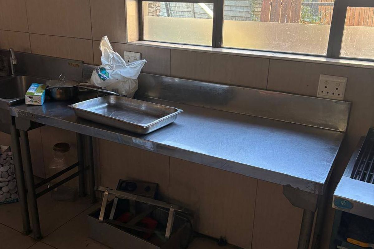 Commercial kitchen prep area featuring stainless steel work surfaces, integrated sink, tiled splashbacks, and functional window for natural light.