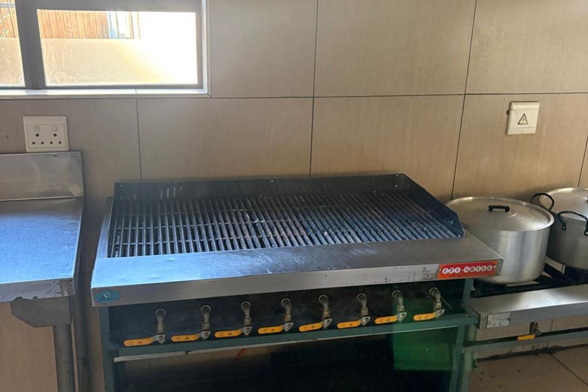 Commercial kitchen area featuring a heavy-duty gas grill, stainless steel prep surfaces, and tiled wall finishes under an extractor hood.