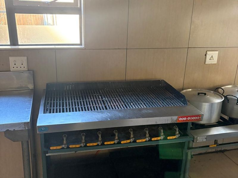 Commercial kitchen area featuring a heavy-duty gas grill, stainless steel prep surfaces, and tiled wall finishes under an extractor hood.