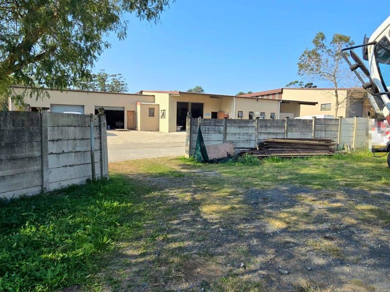 View of the property entrance featuring a precast concrete boundary wall, paved driveway, and industrial warehouse structure on the premises.