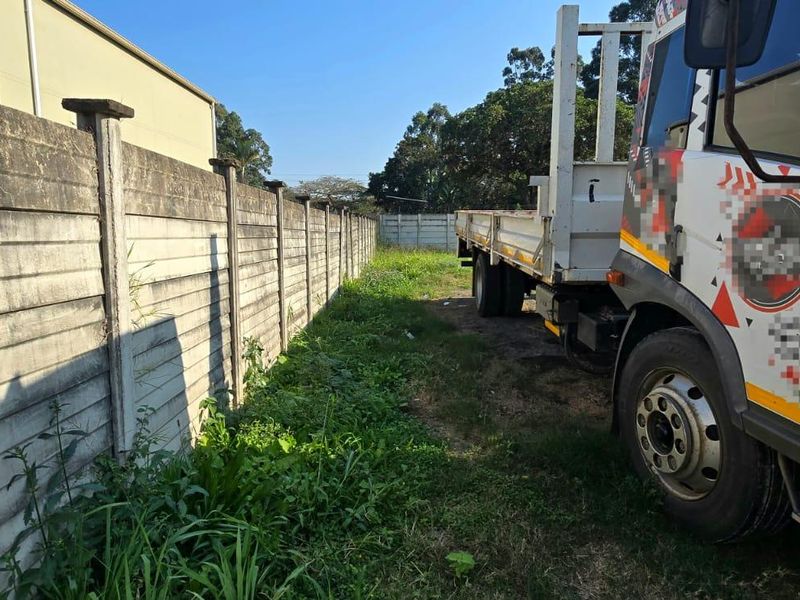 A view of the side or rear boundary of the vacant land, featuring a precast concrete wall and overgrown grass area.