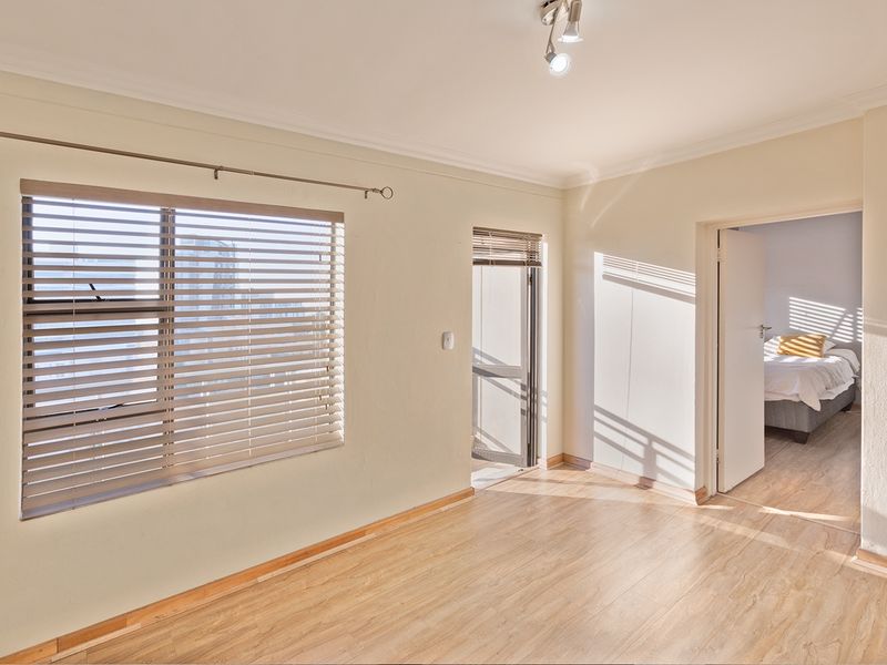 A bright, empty living area featuring light-toned laminate flooring, neutral walls, and venetian blinds, with an adjacent bedroom visible through the doorway.
