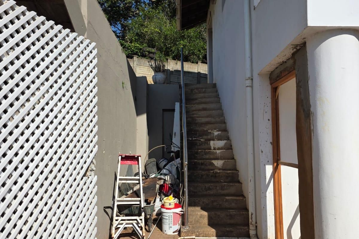Exterior rear area featuring a concrete staircase with a metal handrail, paved walkway, and lattice privacy screen, showing signs of wear and unfinished construction.