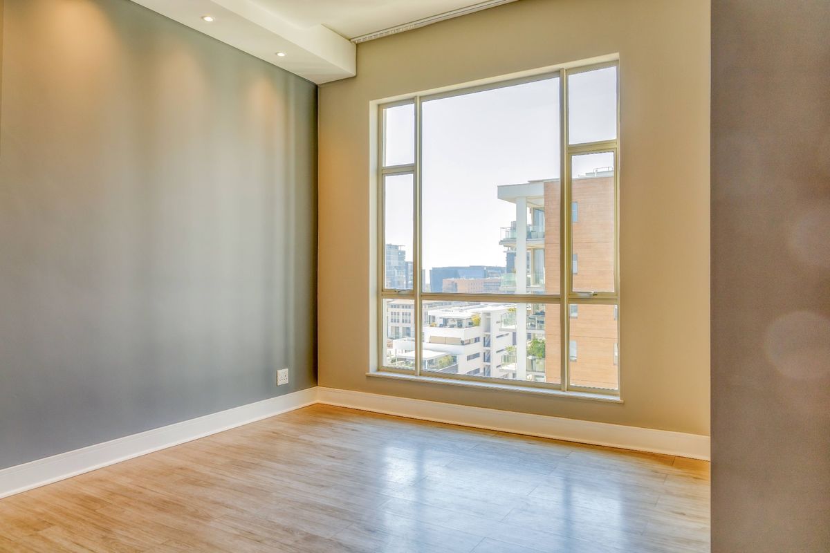 A bright, empty bedroom featuring modern laminate flooring, recessed ceiling lighting, and a large window offering an urban view.