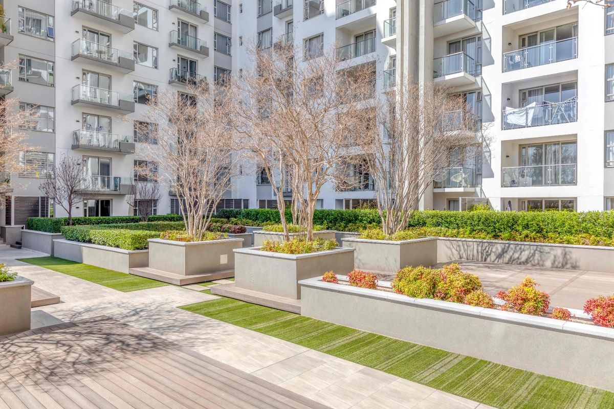 A well-maintained communal courtyard area featuring raised concrete planters, synthetic turf, and paved walkways, surrounded by a modern apartment building with glass-fronted balconies.