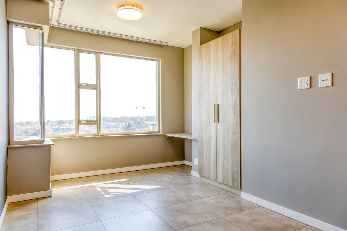 A bright, empty bedroom featuring neutral tiled flooring, a built-in wardrobe, a small integrated desk, and large windows offering natural light.