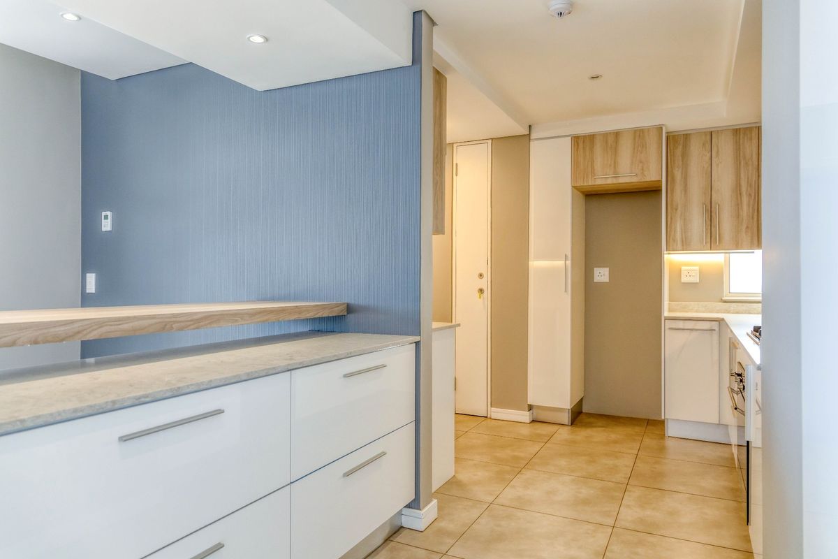 Modern kitchen featuring sleek white cabinetry, stone countertops, a breakfast bar, and warm under-cabinet lighting, finished with neutral tiled flooring.