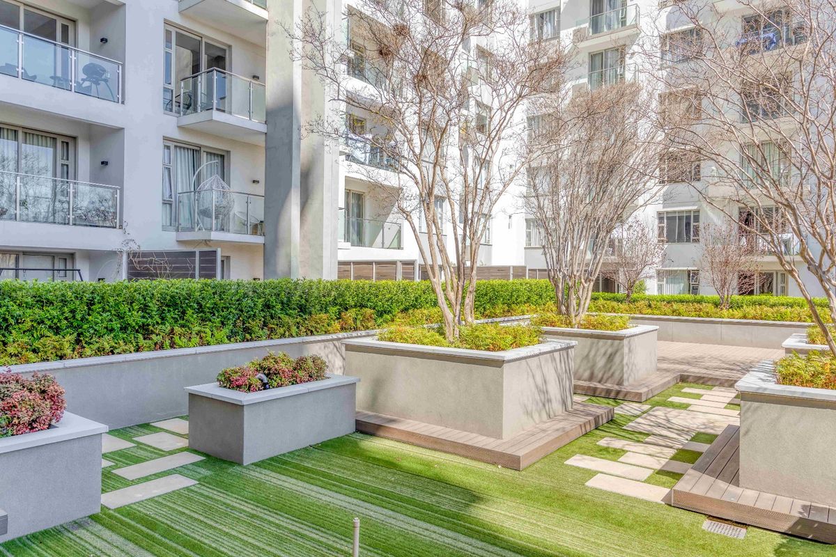A well-maintained communal garden area featuring raised concrete planters, artificial turf, and paved pathways set against a modern apartment building with balconies.