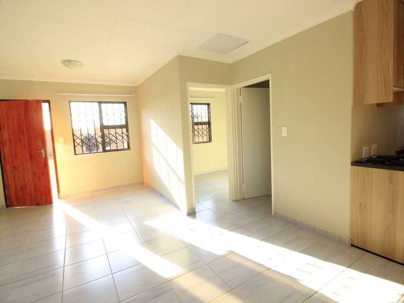 An open-plan living area featuring tiled floors, security gates on windows, and a compact kitchen with built-in cabinetry and a hob.