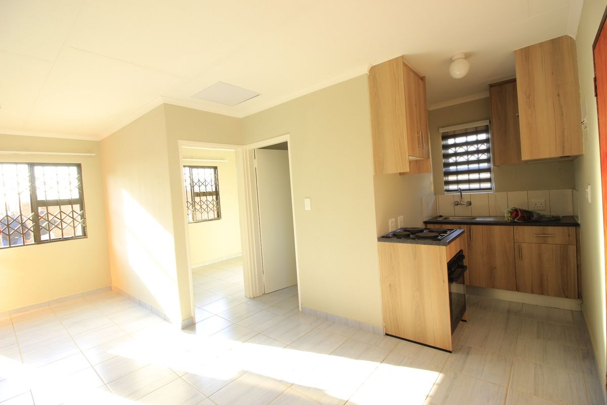 An open-plan living and kitchen area featuring light wood-finish cabinetry, built-in cooking appliances, tiled flooring, and security bars on the windows.