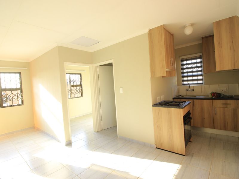 An open-plan living and kitchen area featuring light wood-finish cabinetry, built-in cooking appliances, tiled flooring, and security bars on the windows.