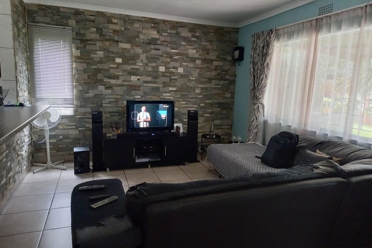 A living room featuring a prominent stone-clad feature wall, ceramic tiled flooring, and large windows with fitted blinds and curtains.