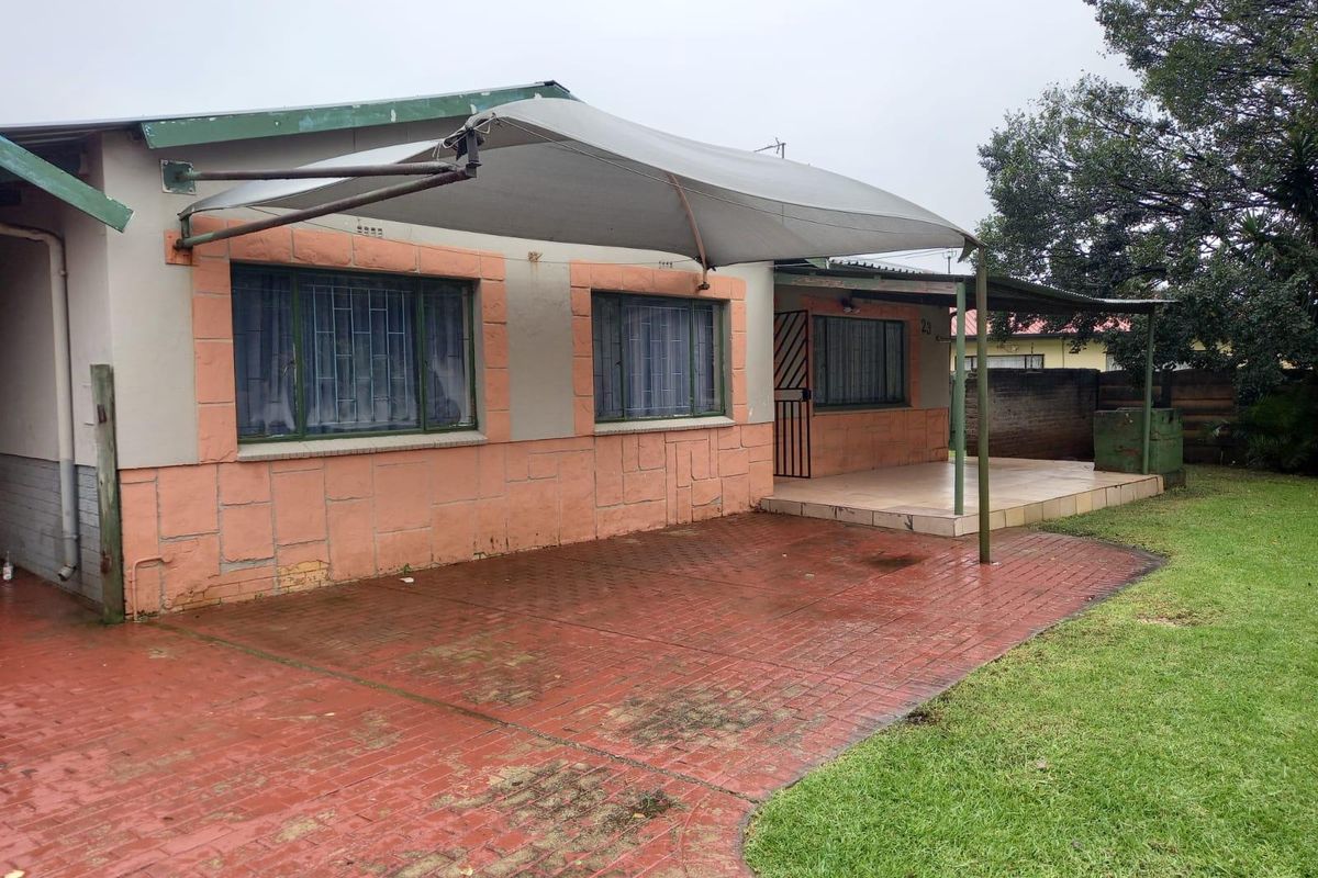 Rear exterior view featuring a brick-paved patio area, a shade sail, and a small lawn, showing signs of weathering and wear.