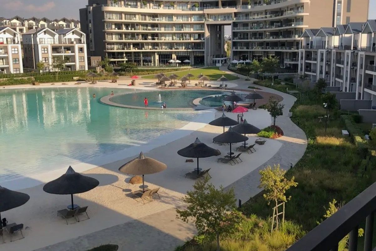 A high-angle view of a large communal swimming pool area within a modern apartment complex, featuring landscaped gardens and paved walkways.