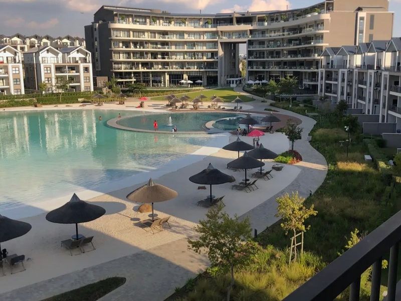 A high-angle view of a large communal swimming pool area within a modern apartment complex, featuring landscaped gardens and paved walkways.