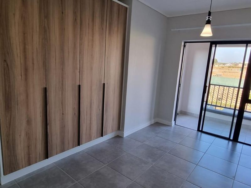 Modern bedroom featuring built-in wood-finish cupboards, neutral tiled flooring, and a sliding glass door providing access to an outdoor balcony area.