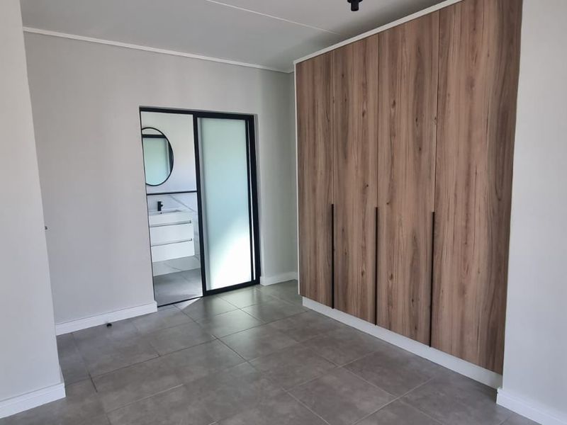 A modern, empty bedroom featuring neutral tiled flooring, built-in wood-finish cupboards, and a sliding door leading to an en-suite bathroom.