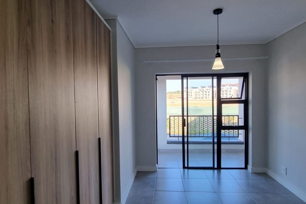 A modern, empty bedroom featuring built-in wooden cupboards, tiled flooring, and a sliding glass door leading to a balcony.