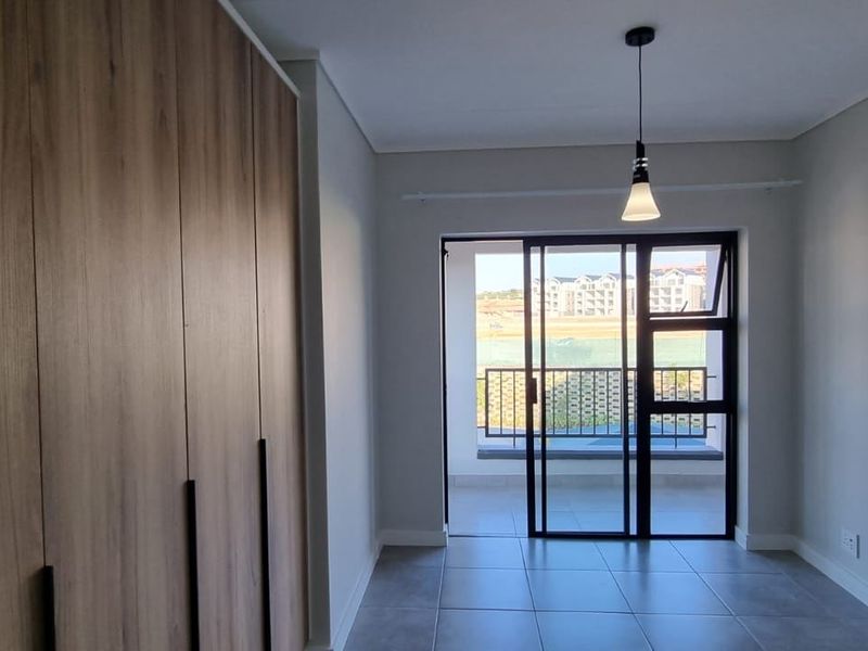 A modern, empty bedroom featuring built-in wooden cupboards, tiled flooring, and a sliding glass door leading to a balcony.