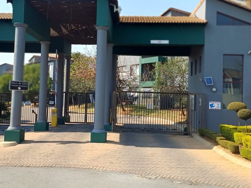 The main entrance and security gatehouse of the apartment complex, featuring paved driveway access and manicured garden hedges.