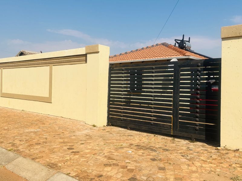 Front view of the property featuring a paved driveway, a secure black metal sliding gate, and a perimeter wall with a solar geyser visible.