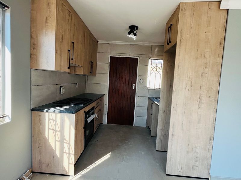 A modern, empty kitchen featuring light wood-finish cabinetry, dark countertops, built-in oven and hob, and tiled backsplash with security bars on the windows.