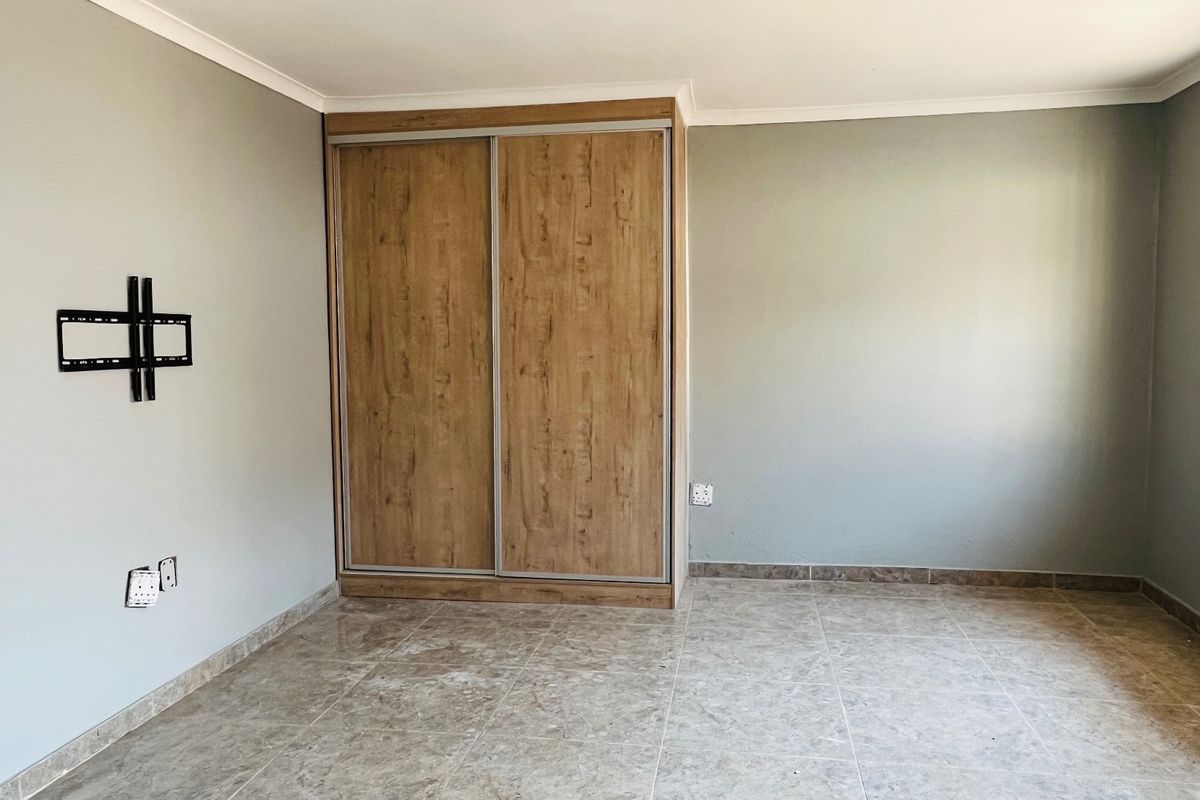 A clean, empty bedroom featuring tiled flooring, a modern wood-finish built-in wardrobe, and security bars on the window.