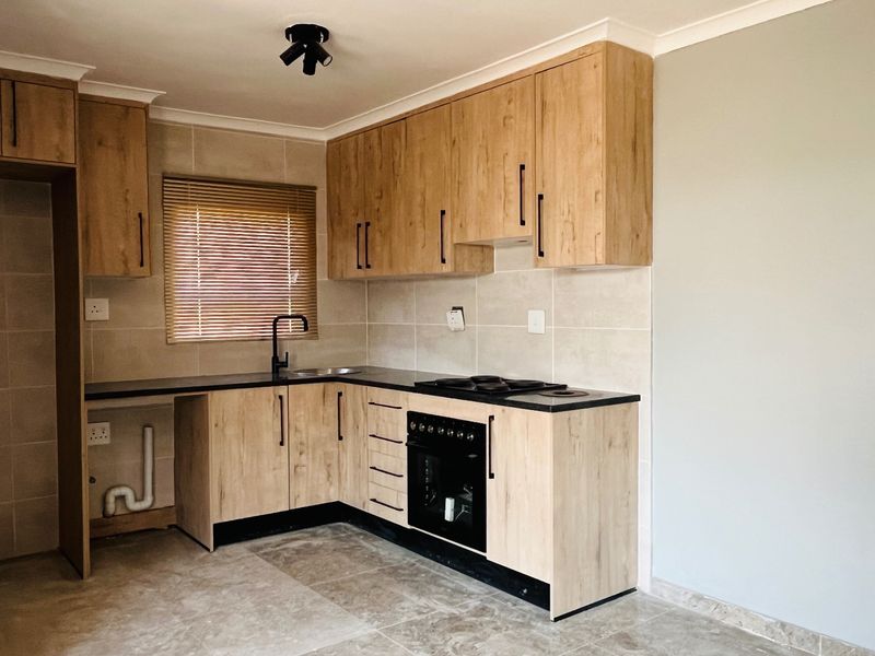 Modern kitchen featuring light wood-look cabinetry, dark countertops, built-in oven and hob, tiled backsplash, and neutral floor tiles in an open-plan layout.