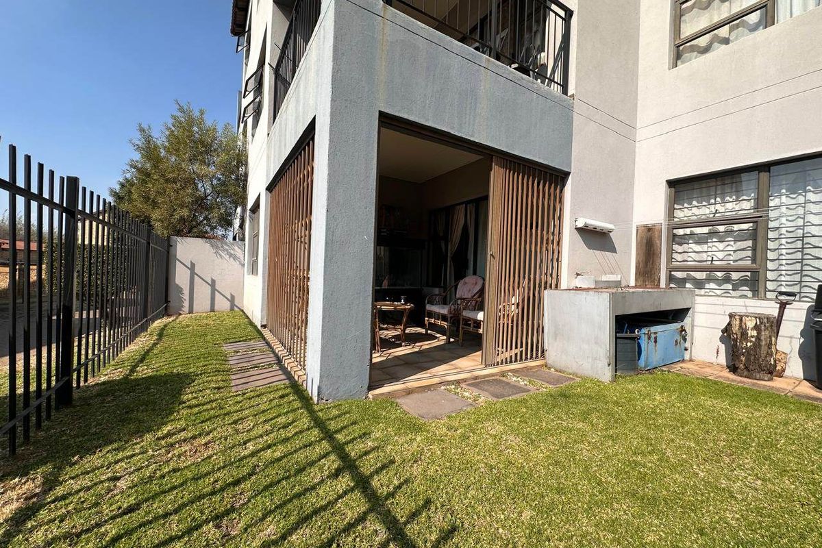 Rear view of the apartment featuring a covered patio with a built-in braai, security gate, and a private garden area with perimeter fencing.