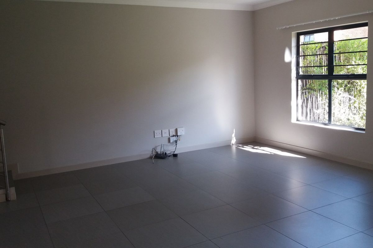 A spacious, empty living area featuring neutral tiled flooring, a staircase with a modern metal railing, and a window with security bars.
