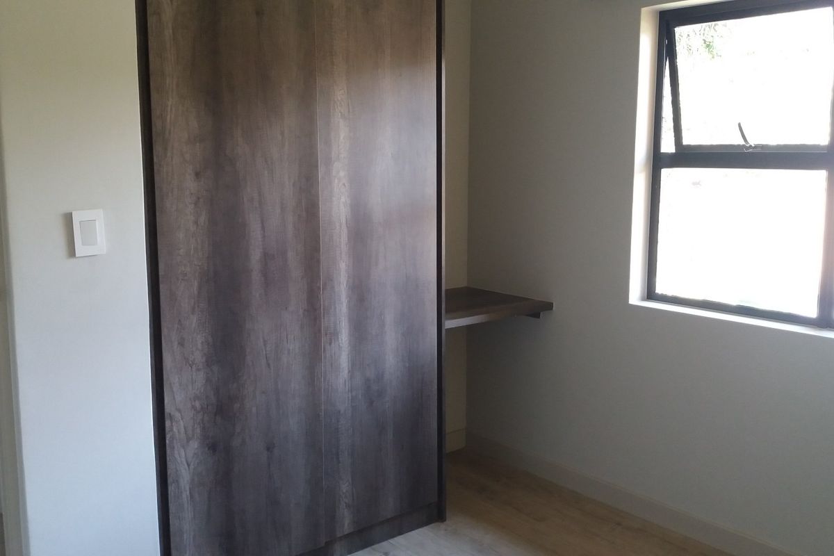 A clean, empty bedroom featuring modern laminate flooring, a built-in dark wood wardrobe, and an integrated desk space under a large window.