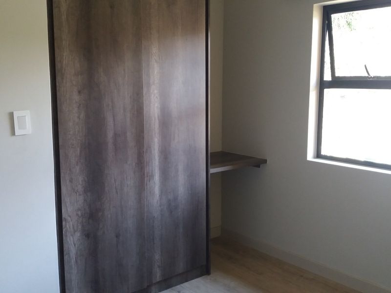 A clean, empty bedroom featuring modern laminate flooring, a built-in dark wood wardrobe, and an integrated desk space under a large window.